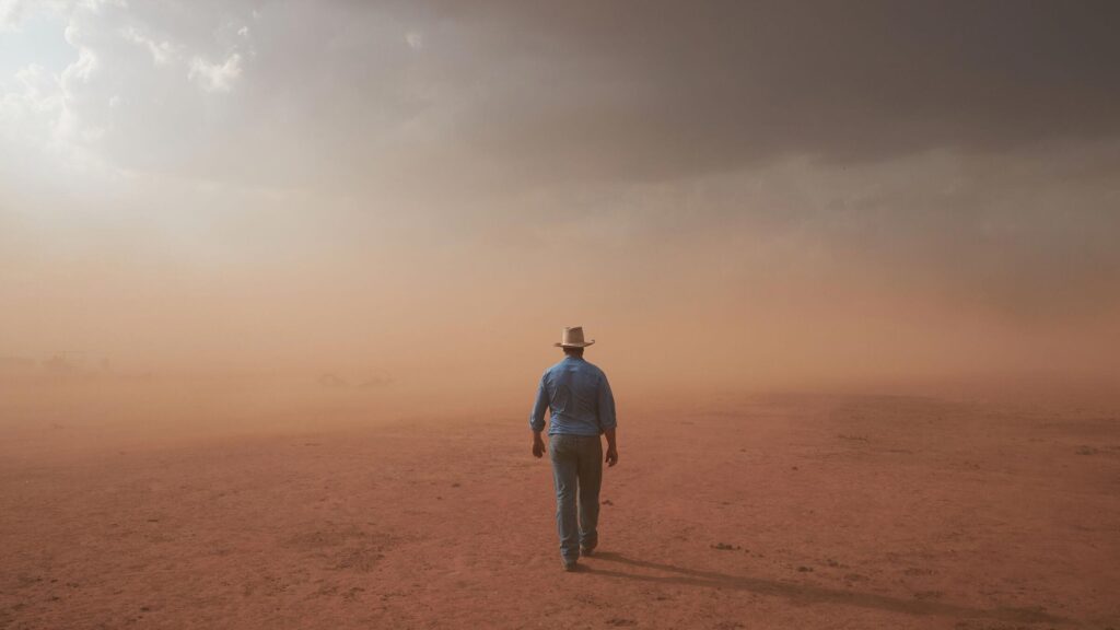 A lone man dressed in outback cattle station riding gear walks into the dusty landscape, dust blowing up ahead of him. He is wearing a blue shirt, blue jeans and cowboy hat. The dusty landscape is red and desolate.